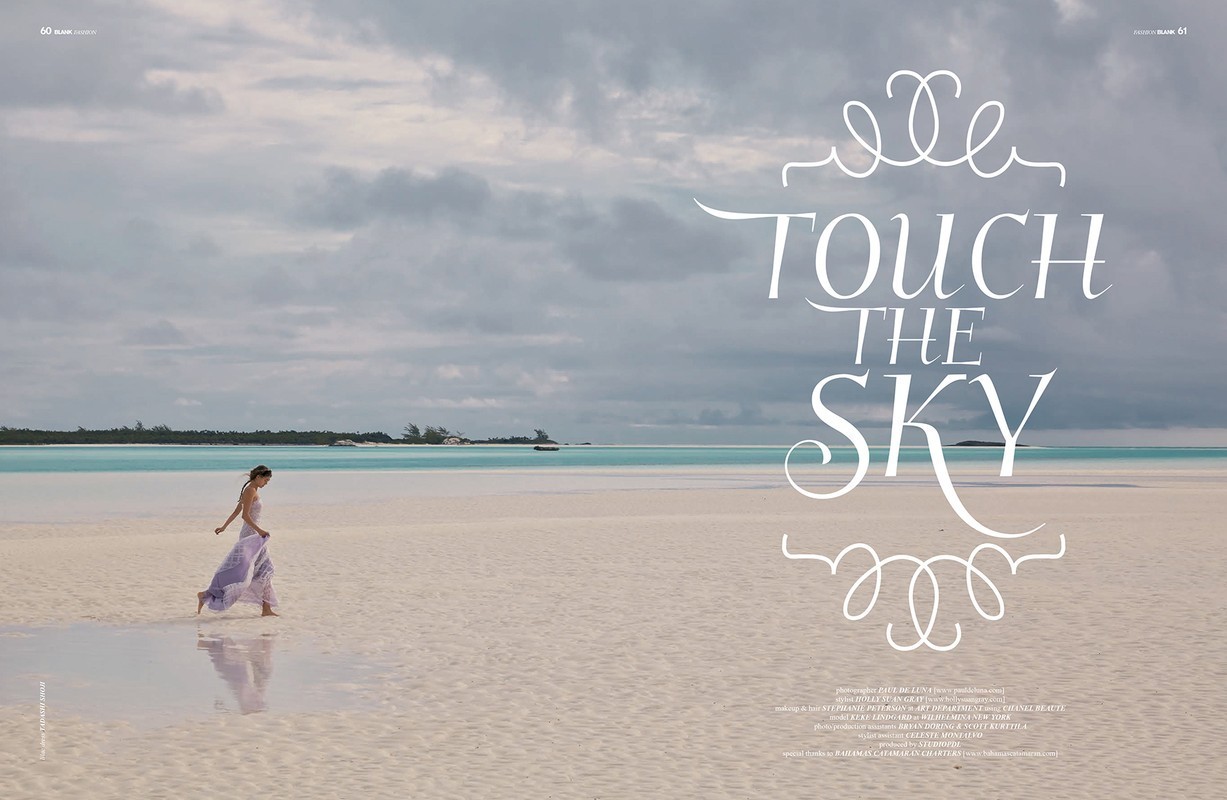 A woman in a light purple dress walks on a bright sand beach next to turquoise water under a cloudy sky, creating a serene and tropical scene.