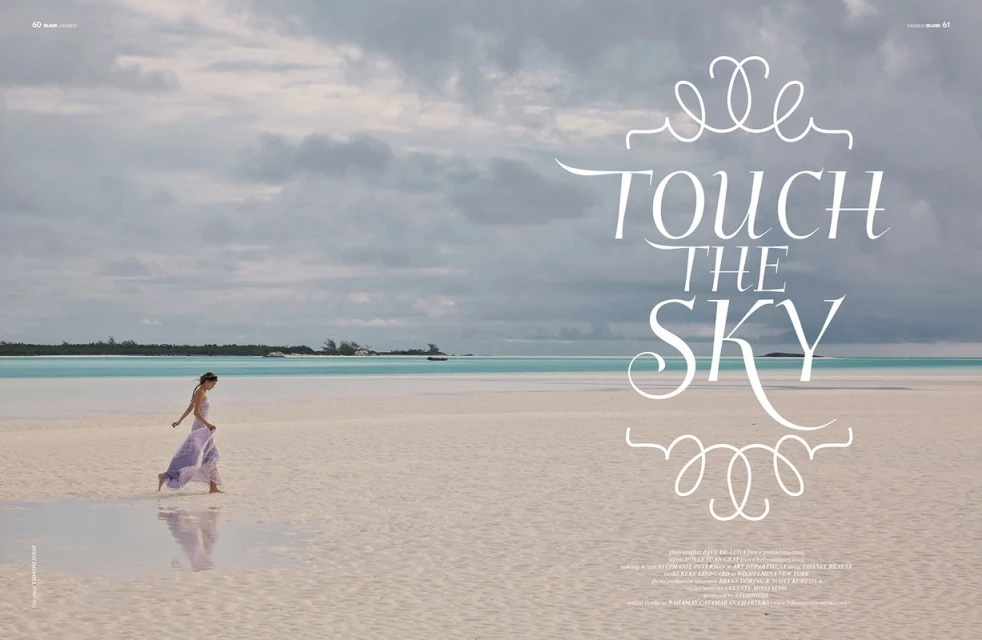 A woman in a light purple dress walks on a bright sand beach next to turquoise water under a cloudy sky, creating a serene and tropical scene.