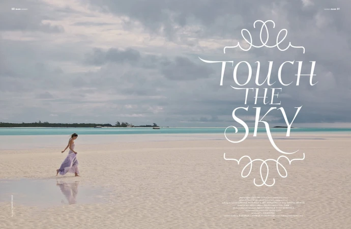A woman in a light purple dress walks on a bright sand beach next to turquoise water under a cloudy sky, creating a serene and tropical scene.