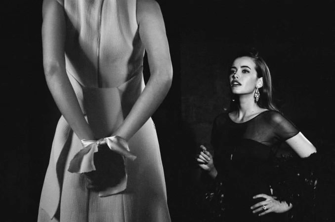 Two women in dresses pose in a black-and-white indoor fashion shot, one with hands tied in bow behind her back, the other with feather boa.