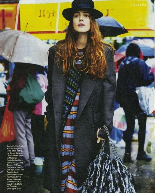 A person wearing a black hat, gray coat, and striped dress stands in a rainy outdoor setting with other people and umbrellas.
