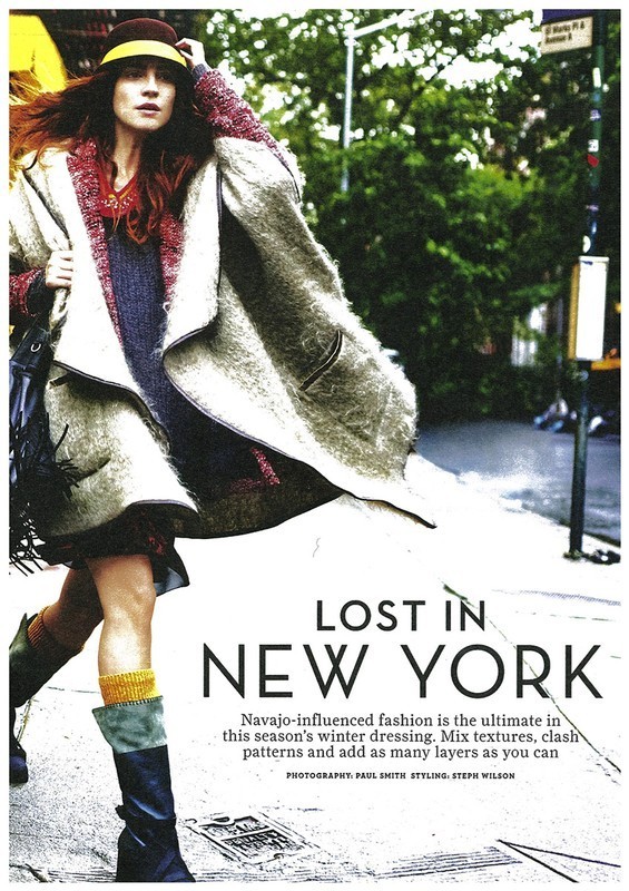 A woman models an oversized coat, brown visor hat, and black boots while walking down a city street with trees in the background.