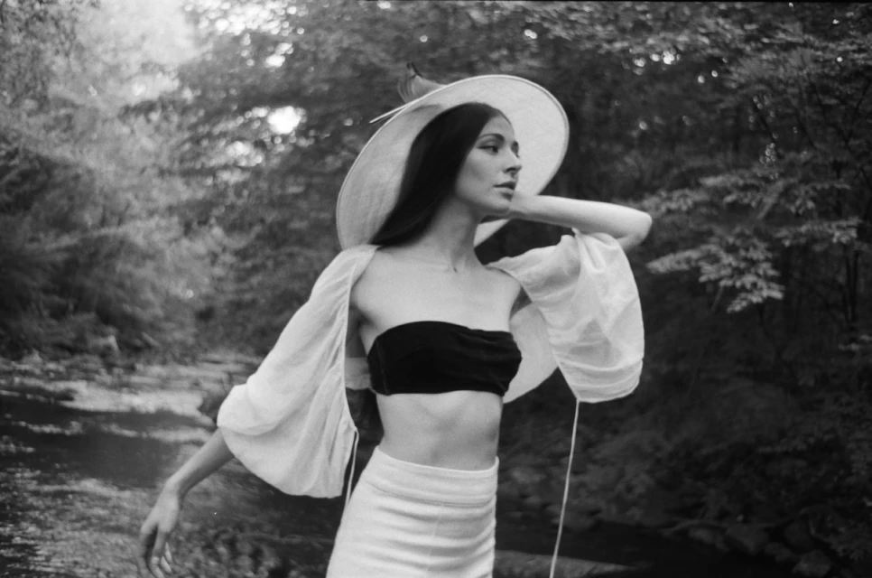 Woman in flowing white sleeves, tube top, and sun hat poses in a lush forest by a creek, creating a captivating, artistic shot.