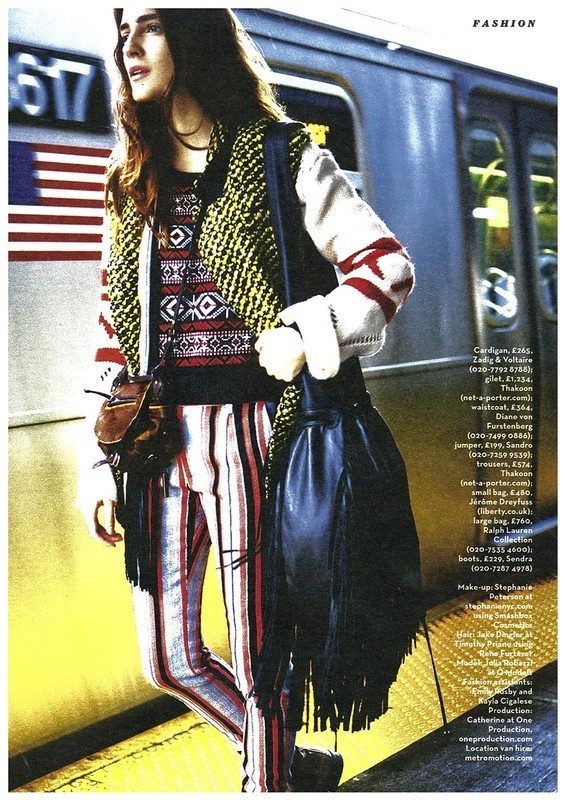 A young woman poses next to a subway train in a fashion editorial, wearing a patterned sweater and striped pants.