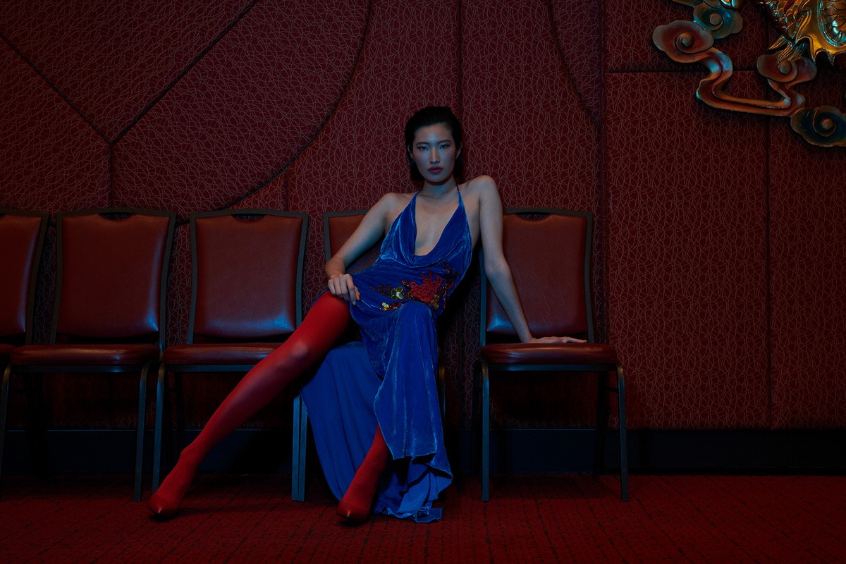 Asian woman in a blue velvet dress and red stockings sits on a chair in a red room, posing for a fashion editorial photo.