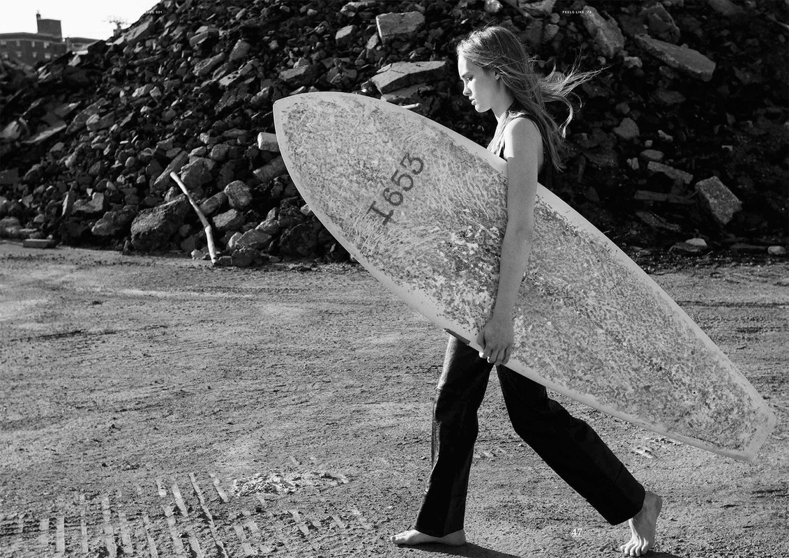 Barefoot woman in black carries surfboard in black and white, rubble background, neutral tone, sandy ground.