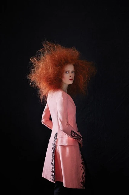 Woman in pink jacket and red hair posing against a black backdrop in a fashion shot, full-length portrait, facing left, eye-level angle.