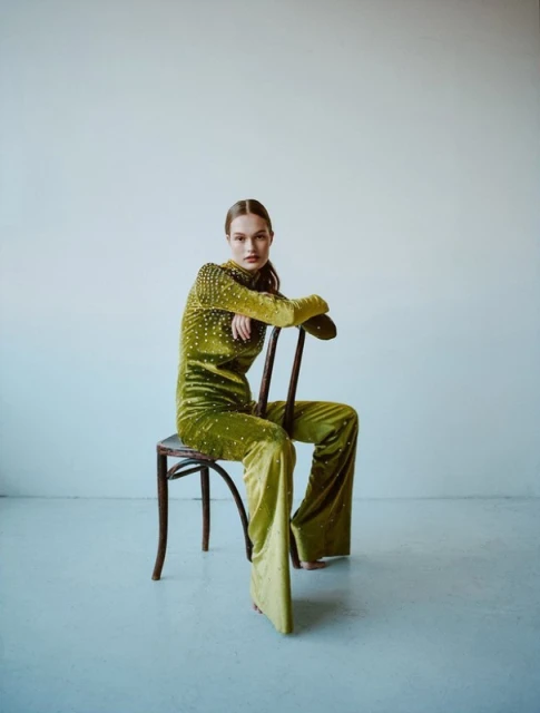A woman in a green jumpsuit with embellishments is sitting on a wooden chair in front of a white wall in a fashion-style image.