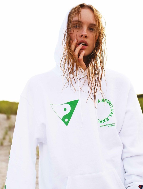 Model in hooded sweatshirt stands outdoors with wet hair, hand to face, against blurred background. White sweatshirt with green graphic design.