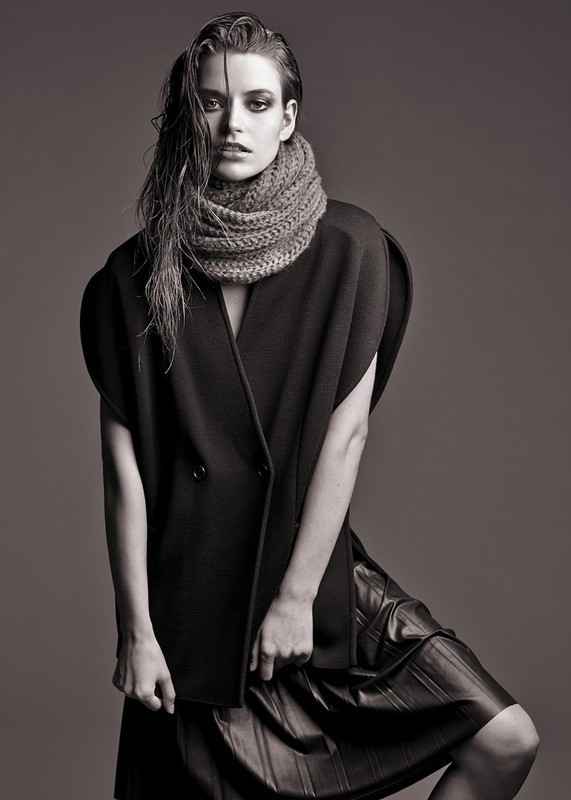 Model in dark sleeveless coat and textured knit scarf with wet-look hair against a grey background