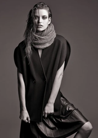 Model in dark sleeveless coat and textured knit scarf with wet-look hair against a grey background