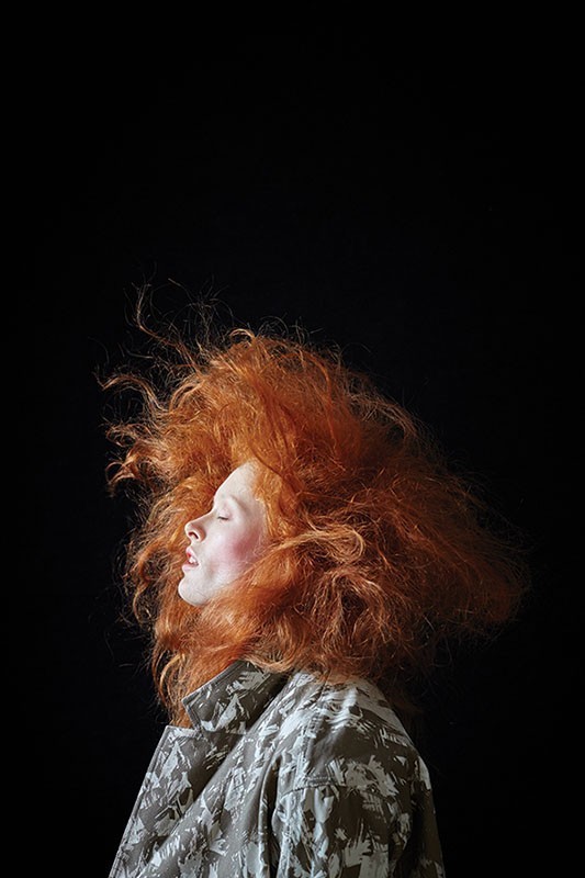 Profile portrait of a woman with wild red hair wearing a patterned jacket against a dark background