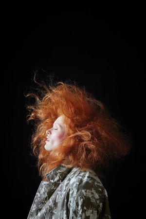 Profile portrait of a woman with wild red hair wearing a patterned jacket against a dark background
