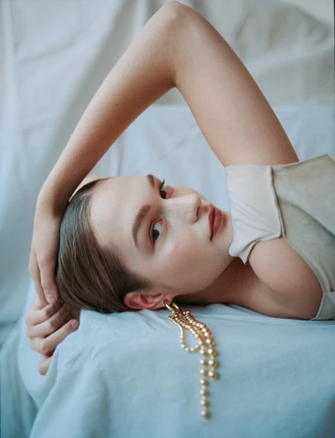 A young woman lies on a light blue surface with a white fabric background, adorned with a large gold earring and wearing a cream-colored top.