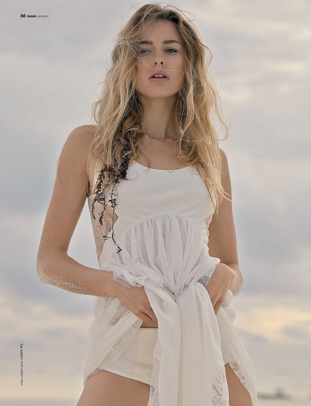 A blonde model poses outdoors on a beach, wearing a short white sundress in a fashion editorial shot. The camera angle is at eye level.
