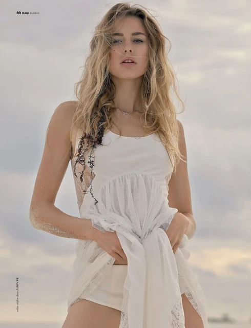 A blonde model poses outdoors on a beach, wearing a short white sundress in a fashion editorial shot. The camera angle is at eye level.