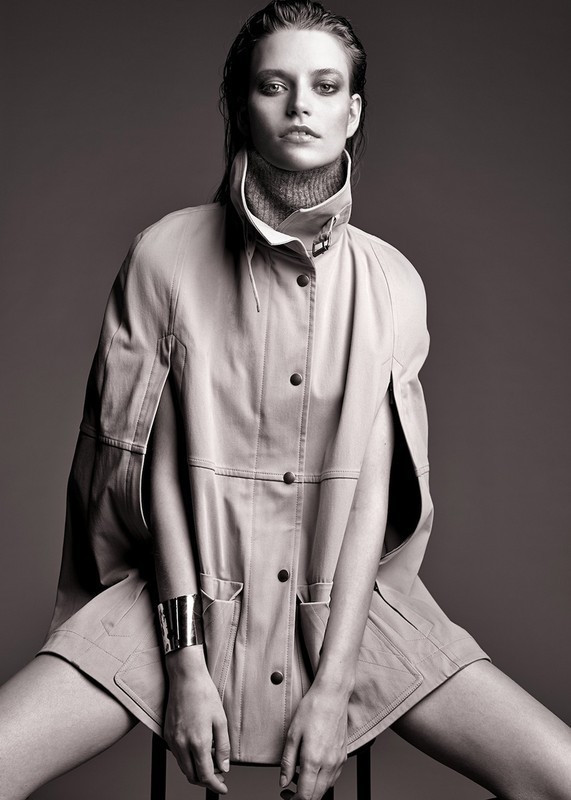 Black and white portrait of a female fashion model wearing a buttoned coat and bracelet while sitting on a stool in a studio.