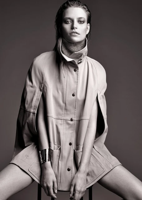 Black and white portrait of a female fashion model wearing a buttoned coat and bracelet while sitting on a stool in a studio.