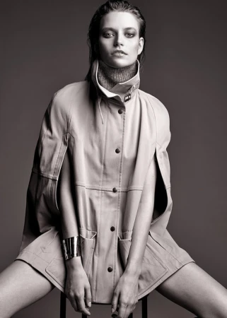 Black and white portrait of a female fashion model wearing a buttoned coat and bracelet while sitting on a stool in a studio.