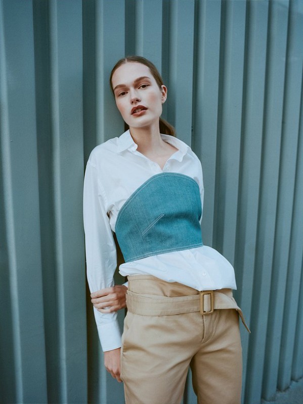 A young woman poses in a white shirt and denim corset with tan pants against a blue corrugated metal wall in natural lighting.