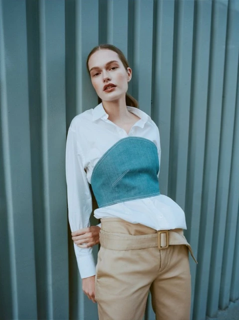 A young woman poses in a white shirt and denim corset with tan pants against a blue corrugated metal wall in natural lighting.