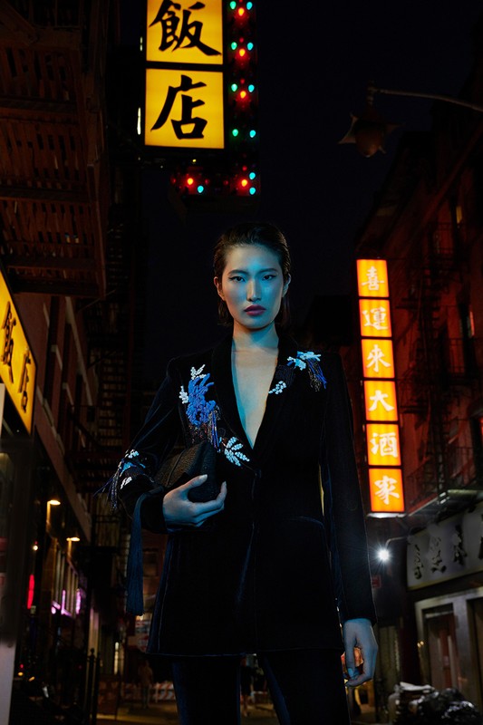 Stylish woman in a dark embroidered velvet blazer stands on a dimly lit city street at night with Chinese restaurant signs.