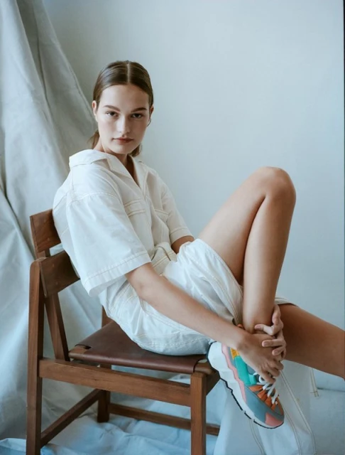 A young woman poses in a short white outfit and colorful running shoes, seated on a wooden chair in a minimalist indoor setting.