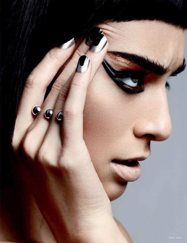 Close-up of a woman with dramatic eye makeup and silver nails, wearing a striking ring with silver orbs, looking off-camera.