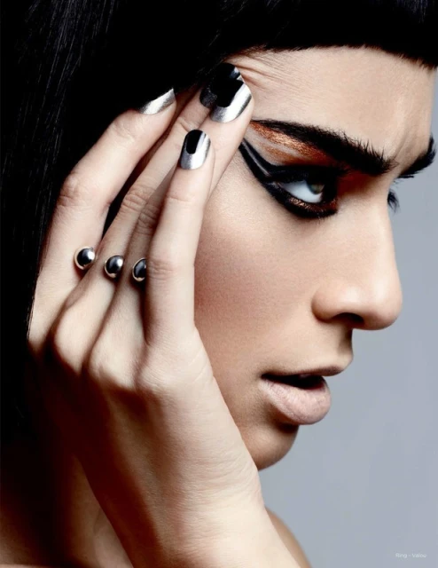 Close-up of a woman with dramatic eye makeup and silver nails, wearing a striking ring with silver orbs, looking off-camera.