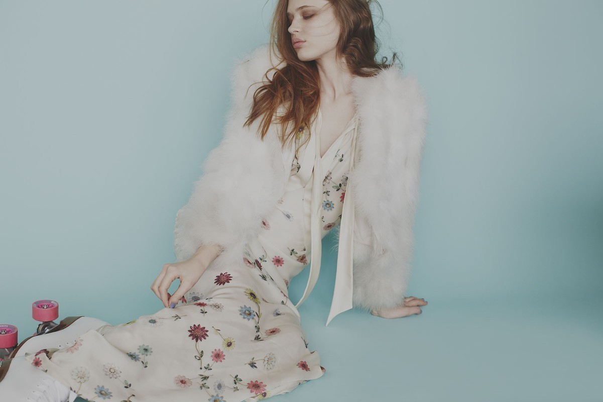Portrait of a young, long-haired, red-haired female model in a floral dress and white coat sitting near pink roller skates on a blue background.