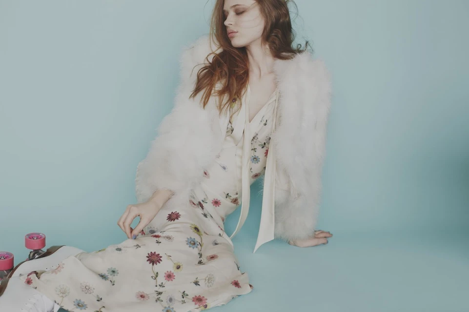 Portrait of a young, long-haired, red-haired female model in a floral dress and white coat sitting near pink roller skates on a blue background.