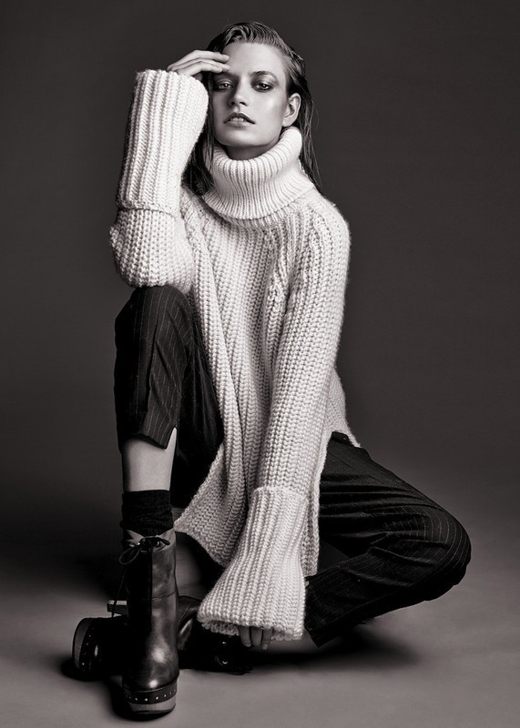 Monochrome portrait of a female fashion model in a knit sweater and pinstripe trousers, seated indoors, with smokey eye makeup.