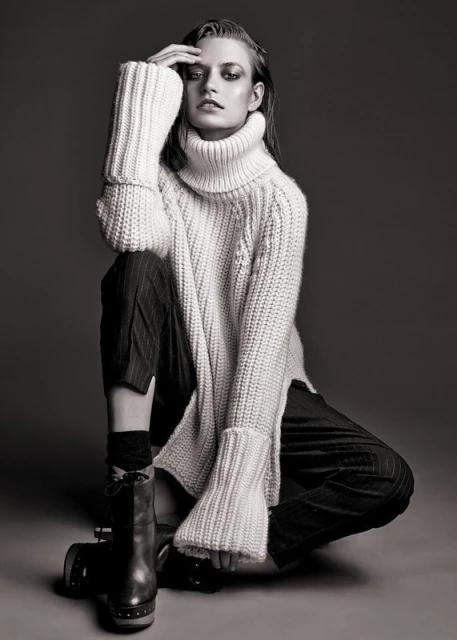 Monochrome portrait of a female fashion model in a knit sweater and pinstripe trousers, seated indoors, with smokey eye makeup.