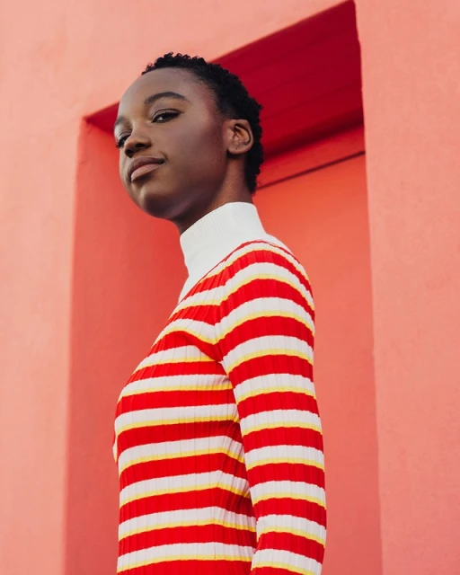 Portrait of a young Black woman in striped sweater against a peach-colored wall, looking slightly upward with a neutral expression.