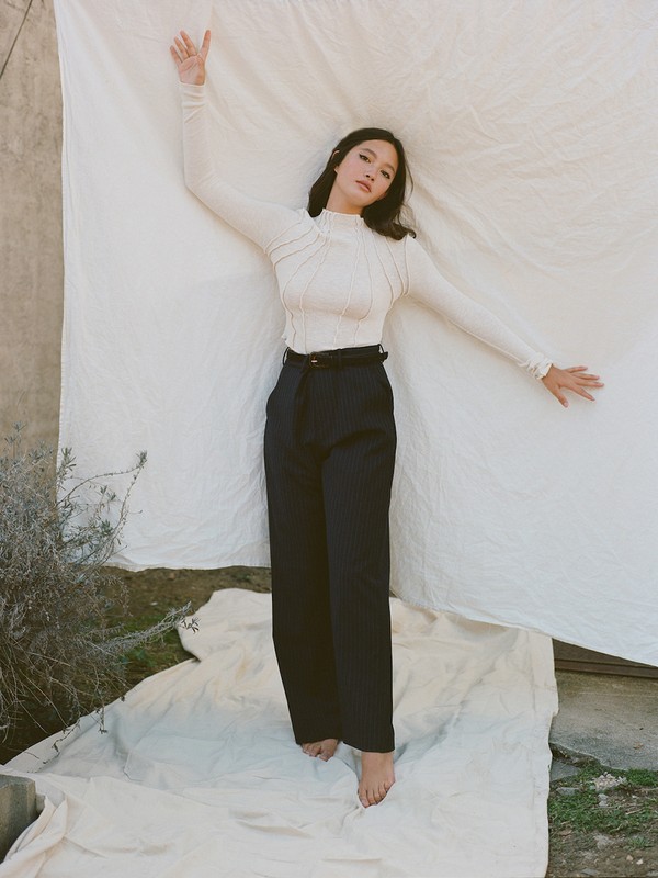 Woman poses in a studio, laying on a wrinkled white cloth, wearing a long-sleeved top and wide-leg black pants, barefoot in natural light.