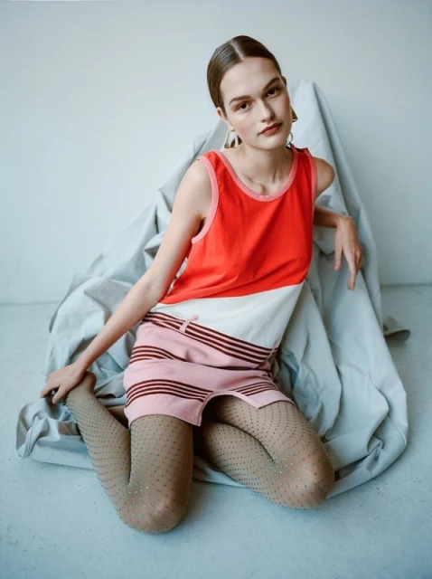 A woman in a color block dress poses in a photo studio, seated on a gray fabric. She has brown hair and is wearing earrings.