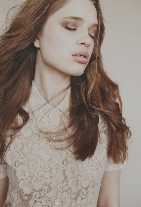 Close-up portrait of a young woman with auburn wavy hair and closed eyes, wearing a floral lace blouse against a neutral background.