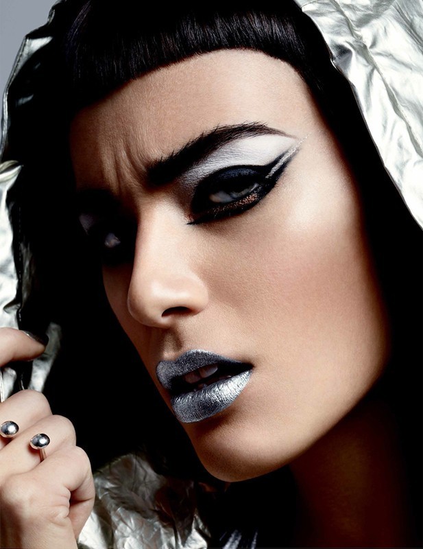 Close-up shot of a woman with bold silver lipstick and white and black eye makeup wearing a hooded silver jacket in a photo studio.