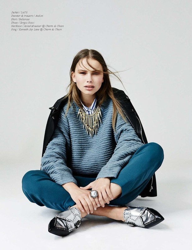 A woman in a blue sweater and teal pants sits cross-legged against a white backdrop, wearing a statement necklace and silver shoes.