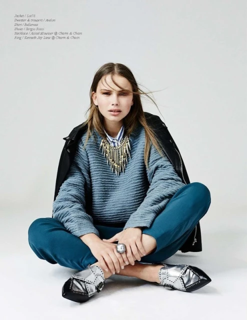 A woman in a blue sweater and teal pants sits cross-legged against a white backdrop, wearing a statement necklace and silver shoes.