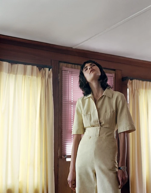 Woman in yellow jumpsuit stands in interior setting with window and curtains, looking upward with neutral facial expression and dark, wavy hair.
