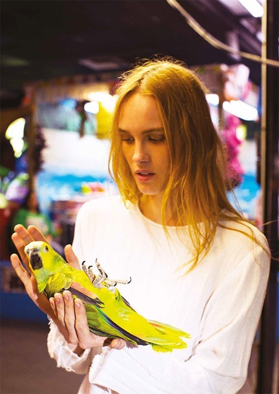 Young woman in a white shirt holding a bright green parrot with yellow head, and pink and blue details on the bird's folded wings.
