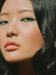 Close-up portrait of an East Asian woman wearing striking teal eyeliner and glossy nude lipstick with dark wavy hair.