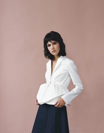 Model with dark hair wearing a white peplum jacket and dark trousers against a dusty rose background.