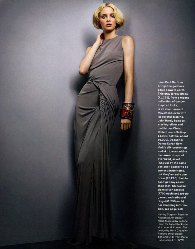 Woman in a long grey gown and large brown cuff stands in front of a grey wall in a fashion studio shot. The light is bright and even.