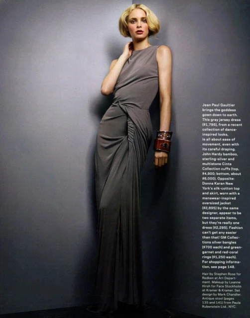 Woman in a long grey gown and large brown cuff stands in front of a grey wall in a fashion studio shot. The light is bright and even.