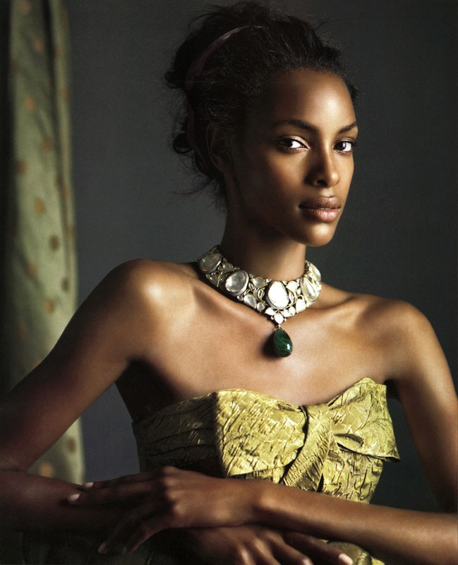 Close-up portrait of a Black woman wearing a green strapless dress and statement necklace, posing indoors against a neutral backdrop.