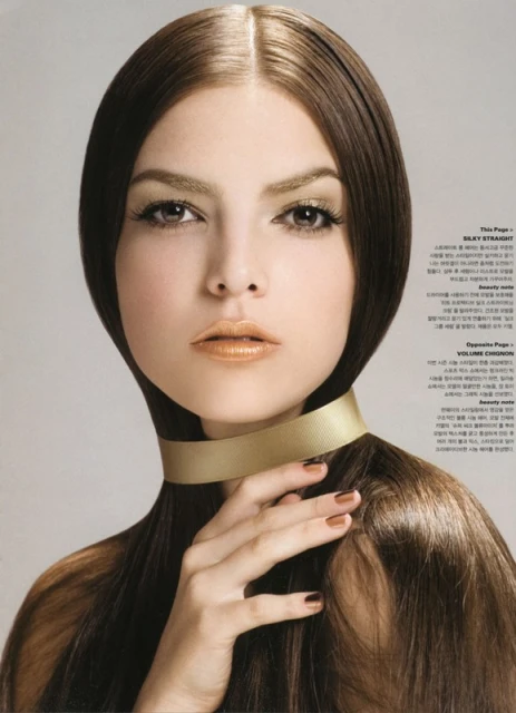 Beauty editorial shot featuring a female model with long brunette hair, gold eyeshadow, and lip color, wearing a gold choker on a grey background.
