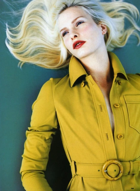 A blonde woman in a golden jumpsuit with a belt poses on a teal background. She is viewed from above and her eyes are closed.