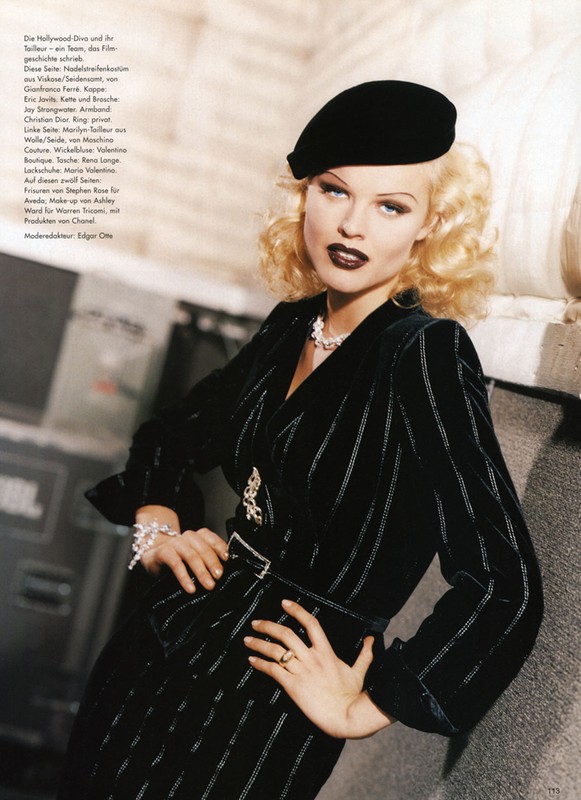 Model in dark pinstripe velvet jacket and black beret leaning against a wall, with styled blonde curls and dark lipstick.